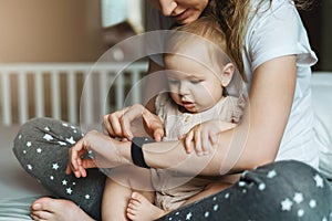 A nine-month-old baby girl with her mother is sitting on the bed and looking at the screen of a smart watch on her mother s hand