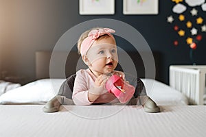 Nine-month-old smiling baby girl in pink bandage sits at white table in highchair and drinks water from baby`s cup
