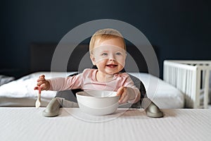 Nine-month-old smiling baby girl sits at white table in highchair and eats herself with spoon from bowl. Blurred background