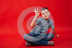 A nine-year-old boy in a shirt in a cage sits against a bright red background and makes a hand gesture. The child shows the symbol