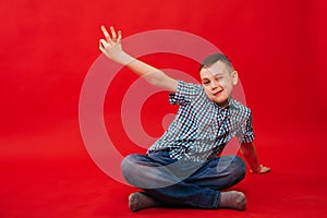 A nine-year-old boy in a shirt in a cage sits against a bright red background and makes a hand gesture. The child shows the symbol
