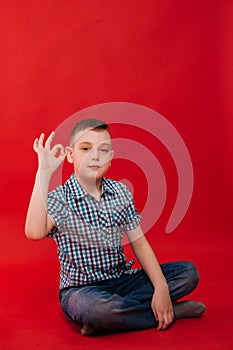 A nine-year-old boy in a shirt in a cage sits against a bright red background and makes a hand gesture. The child shows the symbol