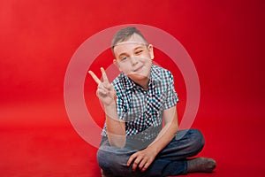 A nine-year-old boy in a shirt in a cage sits on a bright red background and makes a hand gesture. The child shows two fingers