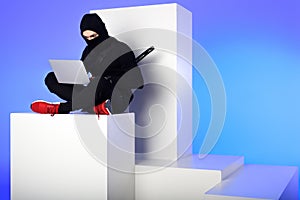 ninja in black clothing using laptop while sitting on white block