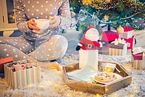 No face woman sitting near xmas tree with open present box and drinking her cocoa with standing near wooden tray with festive