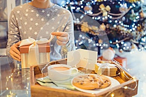 No face young woman opening Christmas present box with blurred wooden tray with festive breakfast. Cocoa and cookies for Santa.