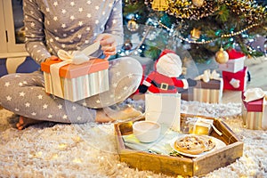 No face young woman opening Christmas present box with blurred wooden tray with festive breakfast. Cocoa and cookies for Santa.