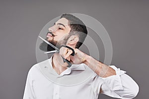 No more beard. Portrait of handsome young man cutting his beard with scissors and looking at camera while standing