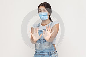 No, please wait, stop. Portrait of scared young brunette woman with medical mask in denim overalls standing and looking at camera