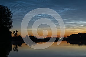 Noctilucent clouds over northwestern Europe, seen from the shore of a lake at night on a calm night