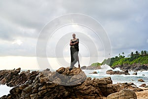 Non-binary black godlike person poses gracefully standing on rocks in ocean. Trans ethnic fashion model in posh dress