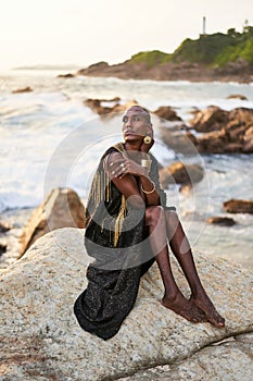 Non-binary black person in luxury dress, golden jewelry on beach rocks in ocean. Trans ethnic fashion model wearing