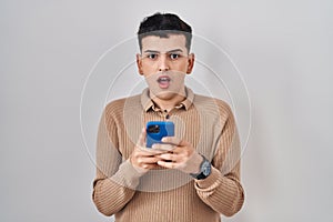 Non binary person using smartphone typing message in shock face, looking skeptical and sarcastic, surprised with open mouth