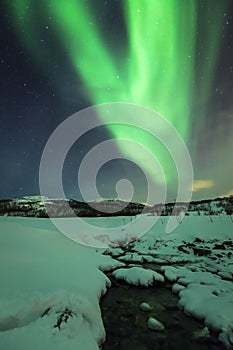 Northern lights Aurora Borealis during night above a water stream in a winter landscape.