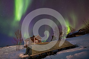 Northern lights over a cabin and a small boat in the snow.