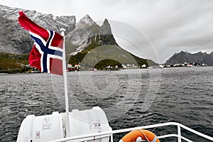 Norwegian flag in the wind on a boat deck on the ocean with mountains and a village in the background on Lofoten Islands in Norway