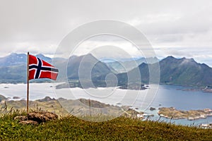 Norwegian national flag waiving in the wind on the top of Nonstinden peak with fjord in the background, Ballstad, Vestvagoy