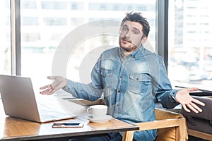 Not sure! Portrait of confused handsome bearded young freelancer in blue jeans shirt are sitting in cafe and working on laptop