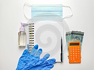 Note book with rubber hand glove,sanitizer bottle,mask,calculator and money on white desk.