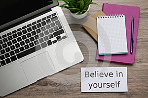 Note with motivational quote Believe in yourself, modern laptop and office stationery on wooden table, flat lay