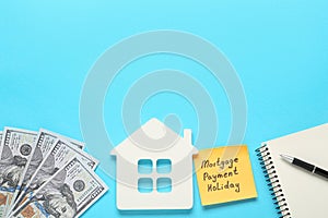 Note with words Mortgage Payment Holiday, house model, notebook and money on light blue background, flat lay. Space for text
