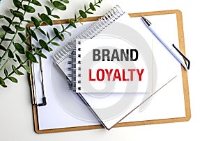 Notebook with brand loyalty text on clipboard surrounded by greenery and office supplies