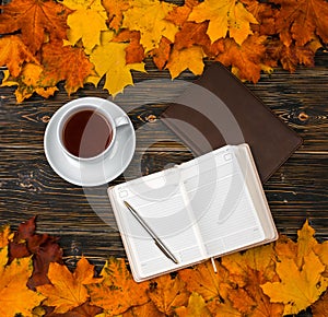 A notebook with an empty space for text, a Cup of tea on a pile of bright autumn leaves against a dark wooden table