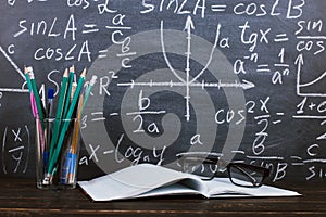 Notebook, glasses and a glass with pencils on chalkboard background with formulas. Teacher's day concept and back to school