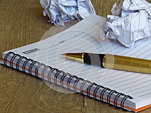 A notebook, a golden metal pen, a pair of black frame glasses and two smashed paper sheet balls on dark wood background.