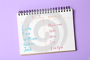 Notebook with lists of baby names on purple background, top view