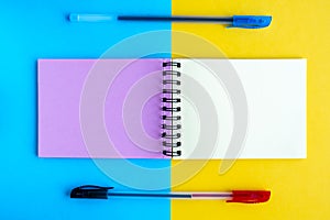 A notebook with multi-colored pages and two ballpoint pens lie on a yellow and blue background. Top view, flat lay, copy space