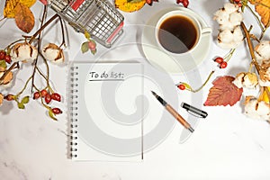 Notebook page wit text To Do List, coffee cup, shopping card and natural autumn decoration on a light marble background, seasonal