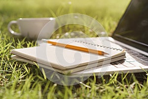 Notebook and pencil are placed on a laptop while the laptop and coffee cup on  green grass In the public park