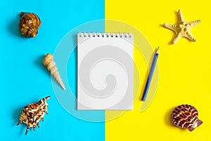 Notebook, pencil with seashells and sea stars on blue and yellow paper background. Mock up, flat lay, copy space, top view. Summer