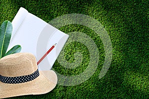 Notebook with pencil and straw hat on green grass background