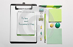 Notebook with phrase Roles And Responsibilities near office supplies on white table, flat lay