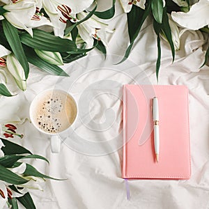 Notebook, present, coffee cup and flowers on the white bed sheet. Good morning and weekend concept in flat lay style