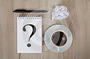 Notebook with question mark, coffee, black pen and crumpled paper. Light colored wooden background