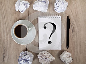 Notebook with question mark, coffee, black pen and crumpled papers. Light colored wooden background
