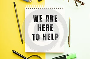 Notebook with text WE ARE HERE TO HELP on office table with office supplies. Yellow color background
