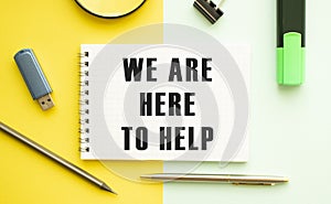 Notebook with text WE ARE HERE TO HELP on office table with office supplies. Yellow color background