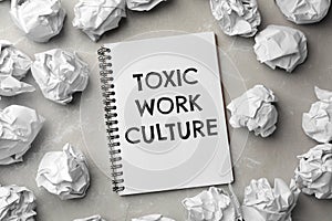Notebook with text Toxic Work Culture and crumpled paper on grey marble table, flat lay