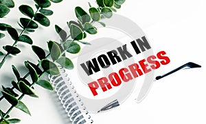 Notebook with text work in progress on white desk with green leaves and pen