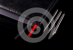 Notebook with three pens over black background