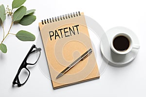 Notebook with word PATENT, pen, cup of coffee, glasses and plant on white table, flat lay