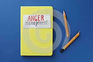 Notebook with words Anger Management and broken pencil on blue background, flat lay