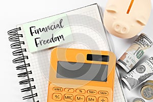Notebook with words Financial Freedom, piggy bank, dollars and calculator on white table, flat lay