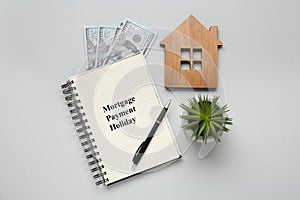 Notebook with words Mortgage Payment Holiday, house model, plant and money on light grey background, flat lay