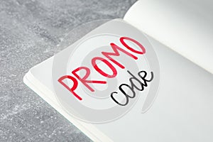 Notebook with words Promo Code on light grey table, closeup