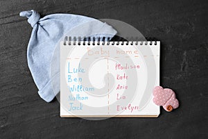 Notebook with written different baby names and child`s accessories on black table, flat lay